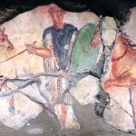 Fresco from the Boukhachkhach (Bou Kachkach) Necropolis (MAR) depicting a soldier, possibly a cavalryman, wearing a blue paenula over a white tunic, holding a green oval shield in his left hand and staff in his right, and with the hilt of a sword visible suspended on his left hip. The horses wear no harnesses or headstalls but do seem to be wearing collars. Late 1st or early 2nd century AD. Image: Dan Diffendale (CC BY-NC-SA 2.0) A soldier stands between two chestnut horses which face him.