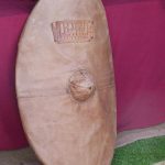 A leather shield cover reconstruction of the Legio I Italica group at Hadrian's Wall Live 2015. Image: MCB A curved oval shield leaned against a wall with a leather cover fitted to it, with patches for the boss and name plate.