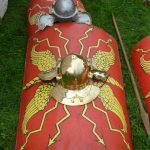 Curved rectangular shield resting on the ground beside a pilum with a helmet placed over the shield boss. Reconstructed by the Ermine Street Guard. Image: MCB Red legionary shield with yellow blazons (wings and thunderbolt motifs) resting on the grass next to a pilum with a brass helmet placed on it.