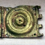 Brass-coloured belt plate with four empty rivet holes, one in each corner.