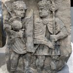 Column base relief from Mainz depicting a legionary with a pilum and a slung helmet and a standard bearer. Image: Heiko Fischer Two infantryman, one on the left with a pilum and curved rectangular shield, with his helmet slung over his neck and pilum. On the right a standard bearer wearing a sagum carries a pole with a single disc on it.