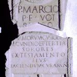Stela of P. Marcius Probus, a custos armorum. He is shown wearing a paenula and carrying a staff of office and a book of writing tablets, both perhaps symbolic of his rank. Image: JCNC Tombstone with a rounded top decorated with a wreath and swags at the top above the inscription. In a rectangular panel at the bottom is a depiction of the deceased and a range of weaponry.