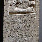 Tombstone of the auxiliary standard bearer, Pancuius, depicting him wearing a mail shirt with shoulder doubling and pteryges, holding a standard on his left side. Image: Hartmann Linge (CC BY) A half-height relief of a soldier is set in an apsidal niche above his inscribed epitaph.