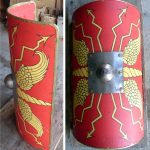Reconstruction of a curved rectangular shield, based on the Dura-Europos original, created by the Ermine Street Guard. Image: MCB A side and front view of a curved rectangular plywood shield painted red with a yellow blazon of wings and thunderbolts. There is a central ferrous boss.