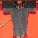 Reconstructed mail shirt with central double-S-shaped fastener in the centre. Image: Álvaro Pérez Vilariño Mail short hanging up on a wooden cross against a red background.