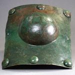 Curved, rectangular copper-alloy shield boss with six of eight domed shield nails in situ. Image: P. Gross Curved rectangular shield umbo with a green patina and with a hemispherical boss in the centre for the hand grip.