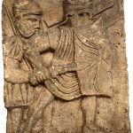 Column base relief from Mainz depicting two legionaries with curved rectangular shields and helmets. The one in the foreground has a drawn sword whilst the man in the background has a shouldered pilum. Image: MCB Two soldiers moving left to right, the one in the foreground and to the right has his weight on his left foot and the sword drawn. That on the right is in the background and has his shield raised.