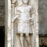 Tombstone of aquilifer L. Sertorius Firmus at Verona. He wears a scale shirt and pteryges with two belts over it, for a sword on his left hip and dagger on his right. He holds an eagle standard in his right hand. Image: MCB Tombstone of an eagle-bearer standing in a niche with his weight on his right leg and not wearing a helmet