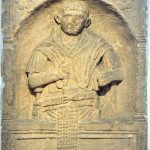 Tombstone of Largennius, a legionary of legio II Augusta. Relief of a soldier shown from the hips upwards but with his apron hanging down over the die of the inscription below. His right hand clutches his paenula, thrown back over his shoulders to reveal his sidearms, whilst his left probably originally held a scroll.