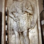 The sculpted relief of Flavoleius Cordus, a pre-Flavian legionary of legio XIIII Gemina from Mainz. It probably pre-dates the invasion of Britain by a few years. He has an oval scutum on his back and holds a shafted weapon in his right hand by means of a throwing strap (amentum). He has sword and dagger belts crossed and wears a tunic with a sagum over it. His boots are depicted with only the sole and central lacing rifge, suggesting the rest of the detail was added in paint. Image: MCB An unarmoured soldier stands in a niche.