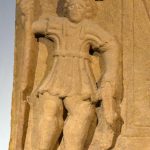 Flanking relief of Mars from an inscription (RIB 1284) from High Rochester suggested by some as depicting an arming doublet (thoracomachus/subarmalis). Image: Carole Raddato (CC BY-SA 2.0) A crude Mars holds a spear in his right hand and rounded shield (resting on the ground) in his left. He wears a garment that consists of vertical folds or quilting.