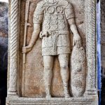 Tombstone of the aquilifer of Gnaeus Musius of legio XIIII Gemia from Mainz. He holds an eagle standard in his right hand and rests his hand on a curved oval shield with a rectangular boss. He wears his awards (torques and phalerae) on a harness of his chest and a sword belt with a horizontally suspended sword. He has fringed pteryges over his tunic and under what is probably mail body armour. The details of his caligae have been carved, rather than mostly painted. He has no dagger or dagger belt. Image: Carole Raddato (CC BY-SA 2.0) Bareheaded soldier standing in a niched tombstone with an inscription detailing his career beneath his depiction.