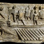 Relief from Praeneste depicting marines on a bireme with a crocodile figurehead. The marines are equipped with oval shields but their equipment shows hellenising tendencies in the shield grips and helmets. Image: Rabax63 (CC BY-SA 4.0) Relief showing the front portion of a bireme moving from right to left, with a double bank of oars, a crocodile figurehead, with a number of marines standing on the deck.